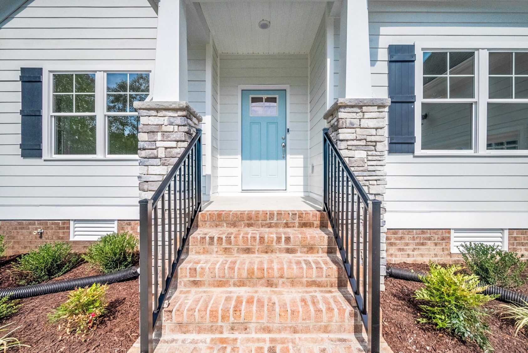 A front entrance of a house with brick steps, a blue front door, white siding, and black metal railings. There are windows on each side of the door and landscaped bushes in front.