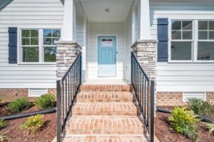 A front entrance of a house with brick steps, a blue front door, white siding, and black metal railings. There are windows on each side of the door and landscaped bushes in front.