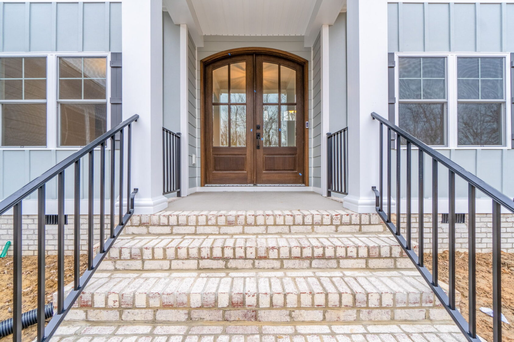 Front entrance of a house with a set of brick steps, black metal railings, and double wooden doors flanked by windows.