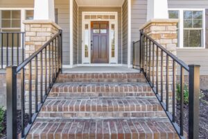 A five-step brick staircase with black railings leads to a wooden front door flanked by sidelights on a gray house with stone accents.