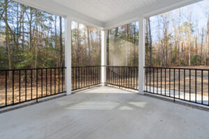 A screened porch with a concrete floor overlooks a wooded area with bare trees. Black railings border the porch, and sunlight casts shadows on the floor.