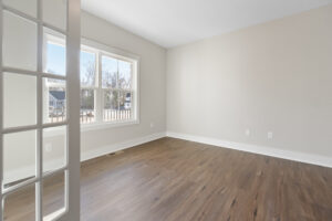 An empty room with light-colored walls, a large window, and hardwood flooring. A glass-paned door is partially open.