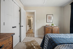 The bedroom in Haley Meadows features elegant wooden furniture, a blue plaid bedspread, open closet and bathroom doors, and soothing beige walls.