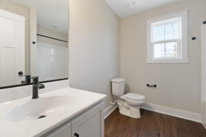 A modern bathroom with a sink, faucet, toilet, and bathtub. The room has a wooden floor, beige walls, and a window with white trim.