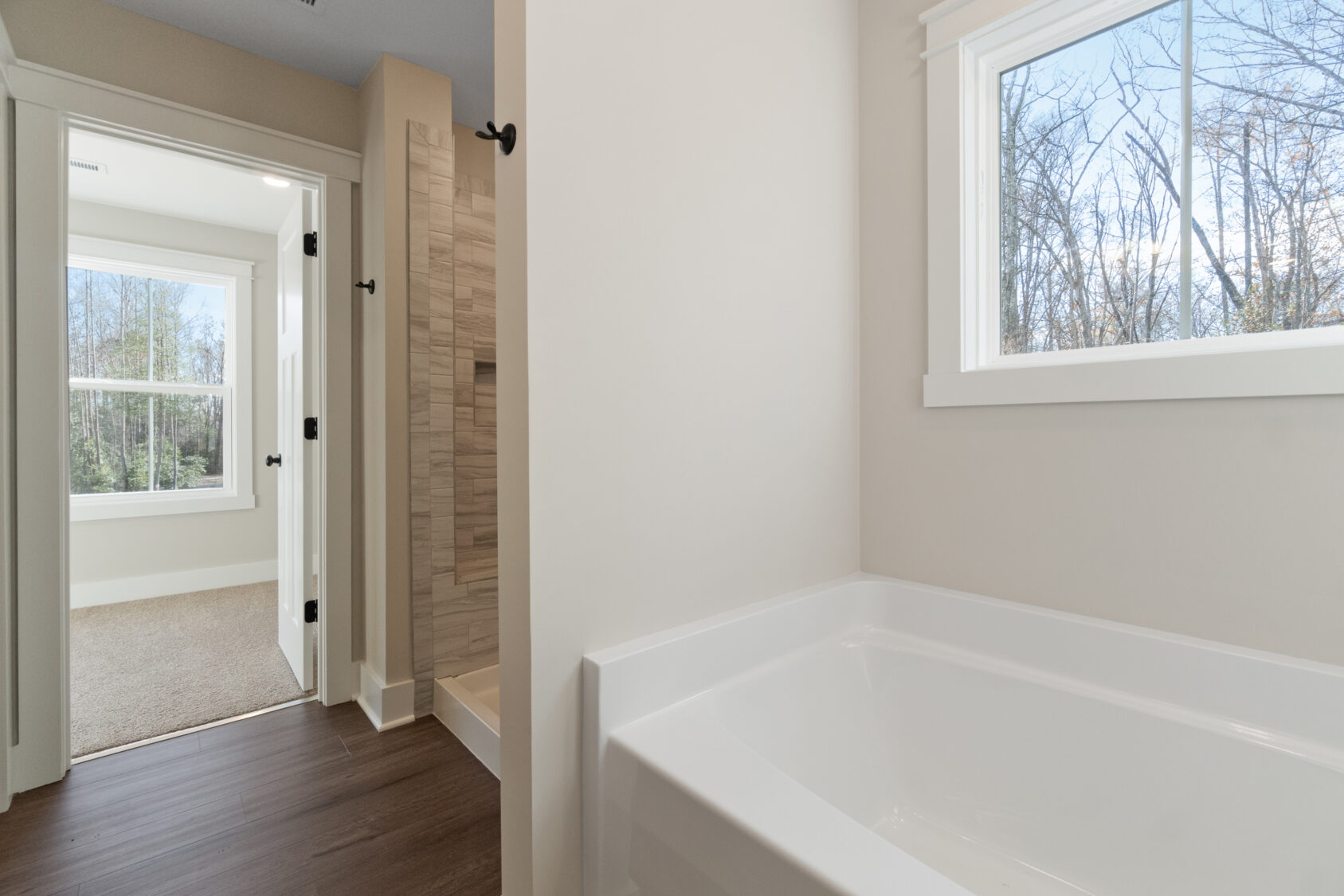 Bathroom with white tub and wooden floor, adjacent to a shower with tiled walls. It leads to a room with carpet and large windows showing a view of trees.