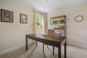 In the Haley Meadows home office, a wooden desk and chair offer a cozy workspace, while dog portraits add charm. Shelves line the walls beside a window that invites natural light, and a classic wall clock keeps time in style.