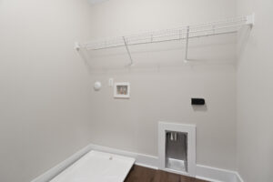 Empty laundry room with a wall-mounted wire shelf, power outlets, and connections for a washer and dryer.