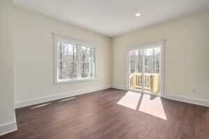 A bright, empty room with light-colored walls, wooden floors, a window, and a glass door leading to a small outdoor deck.