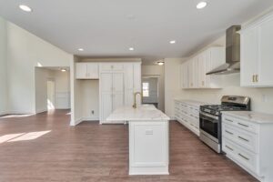 A modern kitchen with white cabinets, stainless steel appliances, a large island with a marble countertop, and wood flooring extending into the adjacent open living area.