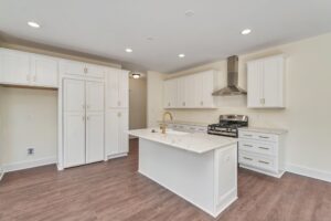 Modern kitchen with white cabinets and an island featuring a marble countertop and gold faucet. Stainless steel stove and range hood are positioned against the wall. Light wood flooring throughout.