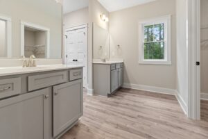 A spacious bathroom with light gray cabinets, dual sinks, wood flooring, and a window with a view of trees.