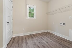 Empty laundry room with light-colored walls, wood flooring, a small window, white door, and wire shelving. Wall cutouts and outlets are present for washer and dryer installation.