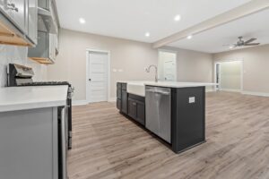 A modern kitchen with wood floors, a large island with a sink, grey cabinets, and stainless steel appliances. Ceiling lights and a fan are visible in the open layout space.