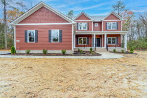 A two-story red house with a porch, surrounded by trees, sits on a sandy yard under Manchesters clear blue sky.