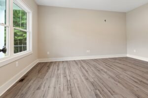 An empty room with light gray wood flooring and beige walls. A large window on the left shows greenery outside.