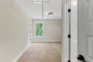 A bright, empty room with a beige carpet, a window showing greenery, and a partially open white door.
