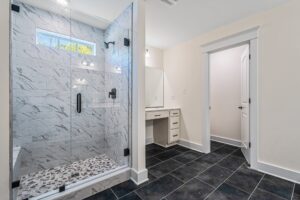 A modern bathroom featuring a glass-enclosed shower with marble tiles and a pebble floor, a vanity with storage, and a door leading to another room. The floor has dark tiles.