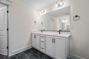 A bathroom featuring a large mirror, double vanity with white cabinets, black hardware, black fixtures, and a white countertop. The room has light-colored walls and dark floor tiles.