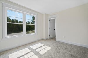 A small, empty room with light-colored walls, a large window with outside view, beige carpet, and an open door leading to another room.