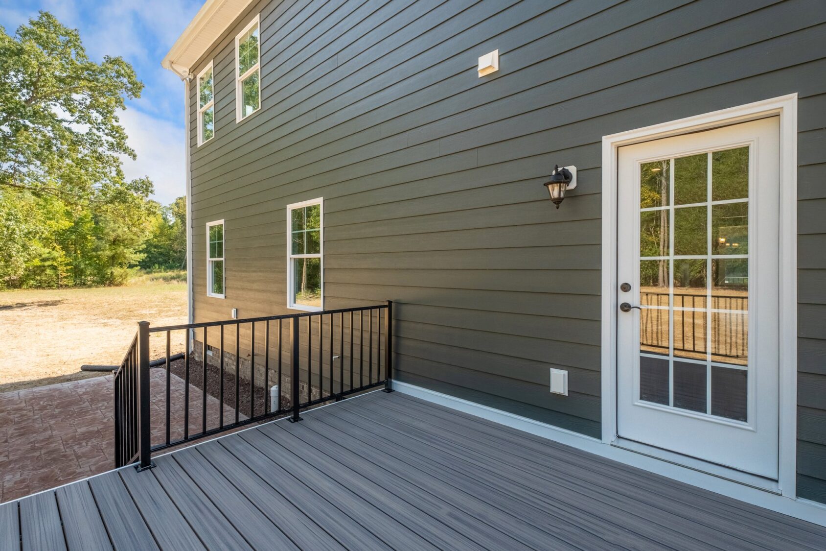 Back view of a house with grey siding, featuring a white door with glass panes, a small outdoor deck with grey flooring and black railing, and several windows. Wooded area is visible in the background.