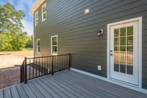 Back view of a house with grey siding, featuring a white door with glass panes, a small outdoor deck with grey flooring and black railing, and several windows. Wooded area is visible in the background.
