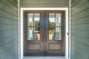 A close-up view of double front doors with glass panes and dark wooden frames, set within a house with green siding.