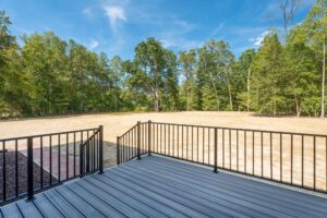 A deck with metal railings overlooks a vast, partly grassy yard with trees in the background under a clear blue sky.
