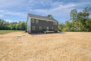 Gray two-story house with a back porch, set in a yard with sparse grass. Surrounded by trees under a partly cloudy sky.