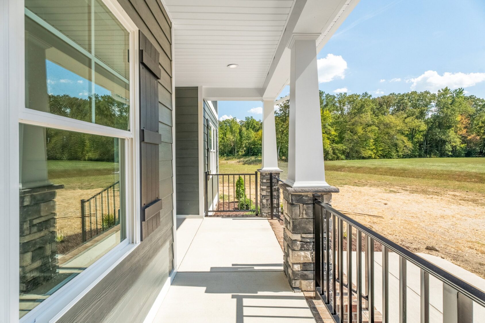 A covered porch with white pillars and a railing overlooks a grassy field. The porch is attached to a house with gray siding and large windows.