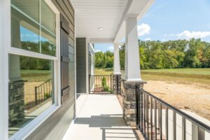 A covered porch with white pillars and a railing overlooks a grassy field. The porch is attached to a house with gray siding and large windows.