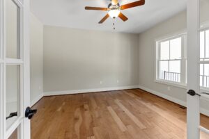 Empty room with wooden floor, ceiling fan, beige walls, large windows, and glass-paned door.