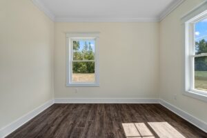 An empty room with light beige walls, two windows showing an outdoor view, and dark brown hardwood floors.