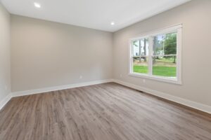 An empty room with light gray walls, a large window, and wood flooring. Natural light enters through the window, overlooking greenery outside. Ceiling lights are on.