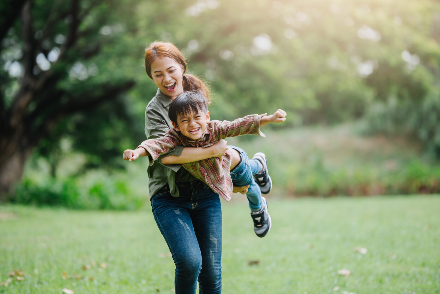A woman twirls a smiling child in the air while standing on a grassy field with trees in the background.