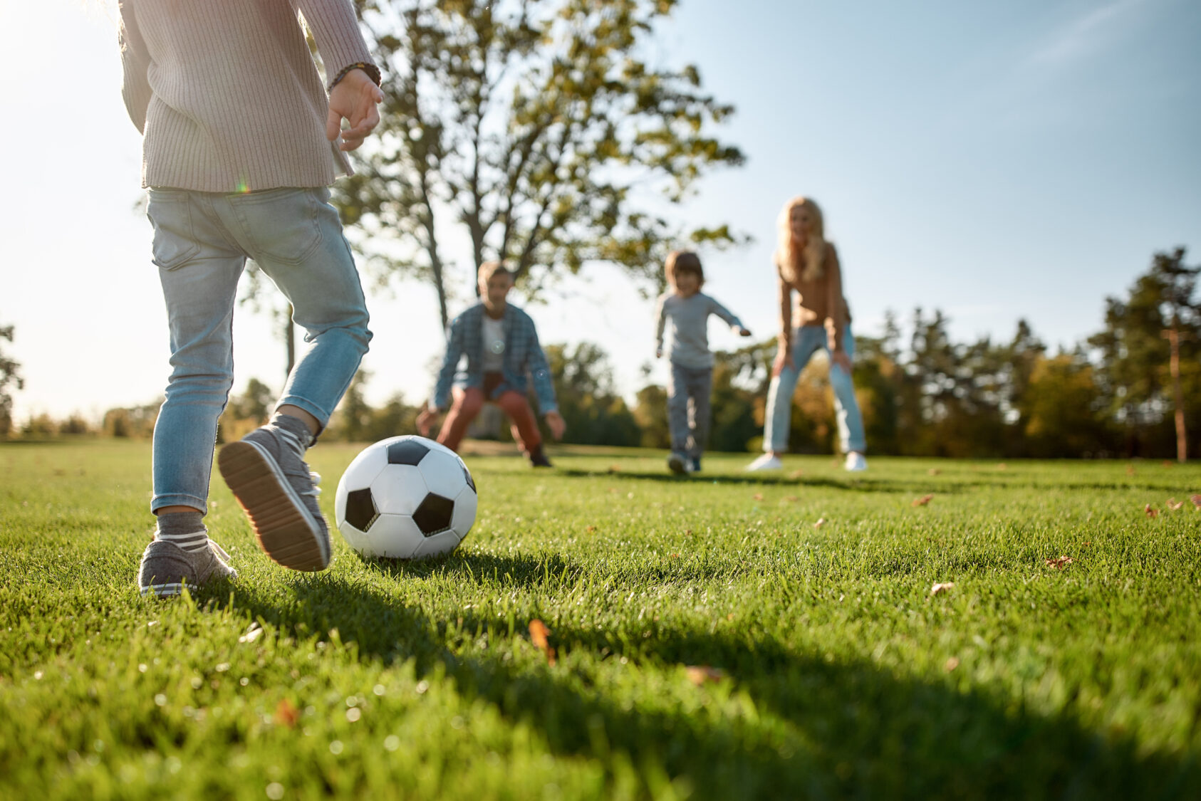 A child kicks a soccer ball across the grassy field of Poplar Village, with three people enjoying the sunny day in the background.