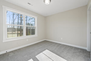 The bright, empty room on Lot 19 features beige walls, a large window overlooking Autumn Hill, gray carpet, and an overhead light.