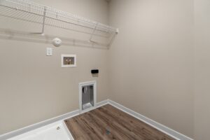 This image shows an empty laundry room with beige walls, a wire shelf, electrical outlets, a wall vent, and wood-patterned vinyl flooring.