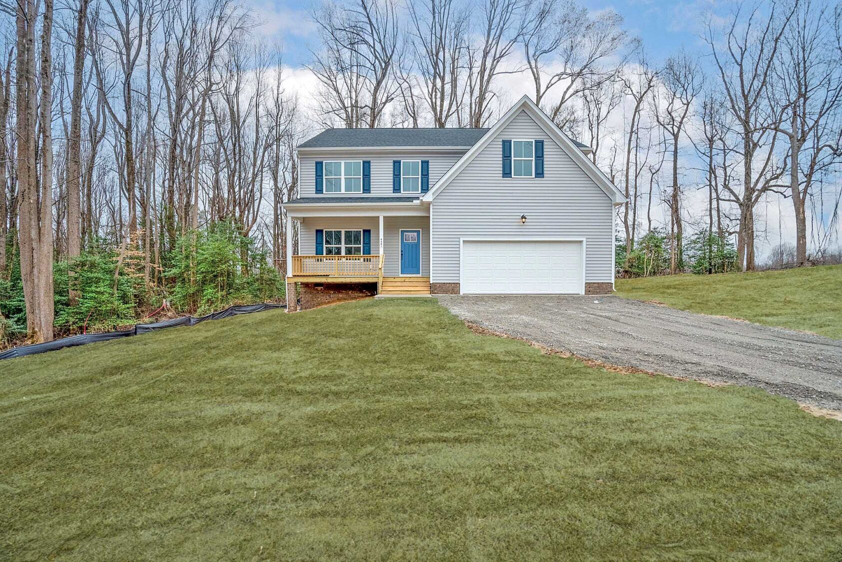 A two-story house with a gray exterior and attached two-car garage, situated on a newly landscaped lawn with surrounding trees in the background.