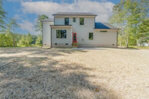 The two-story house in Scotchtown Estates features a red front door, nestled amidst an open yard and trees under a partly cloudy sky.