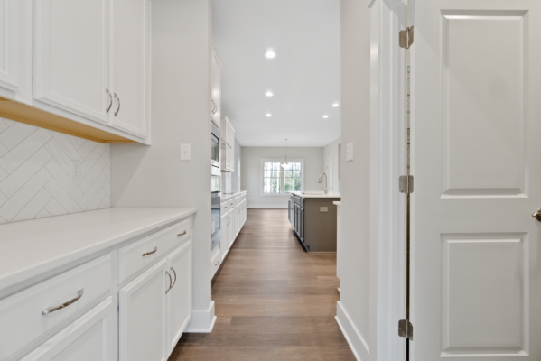 Modern kitchen with white cabinets, herringbone backsplash, and wood flooring in a new home, leading to a room with large windows.