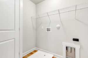 Empty laundry room in Poplar Villages Lot 22 with white walls, wire shelf, and hookups on a wooden floor.