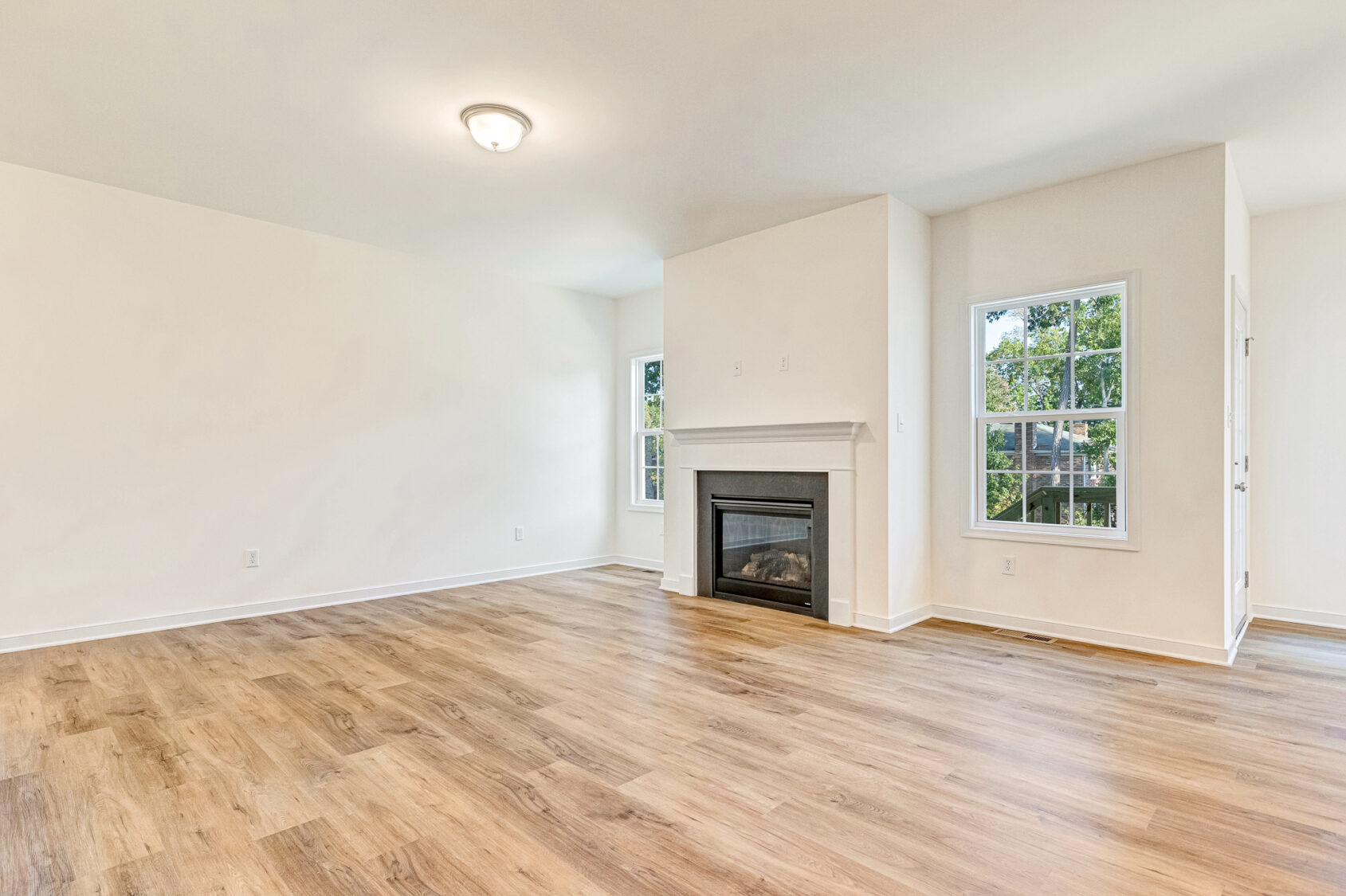 Bright living room in Poplar Village Lot 29 with light wood floors, white walls, fireplace, and large windows.