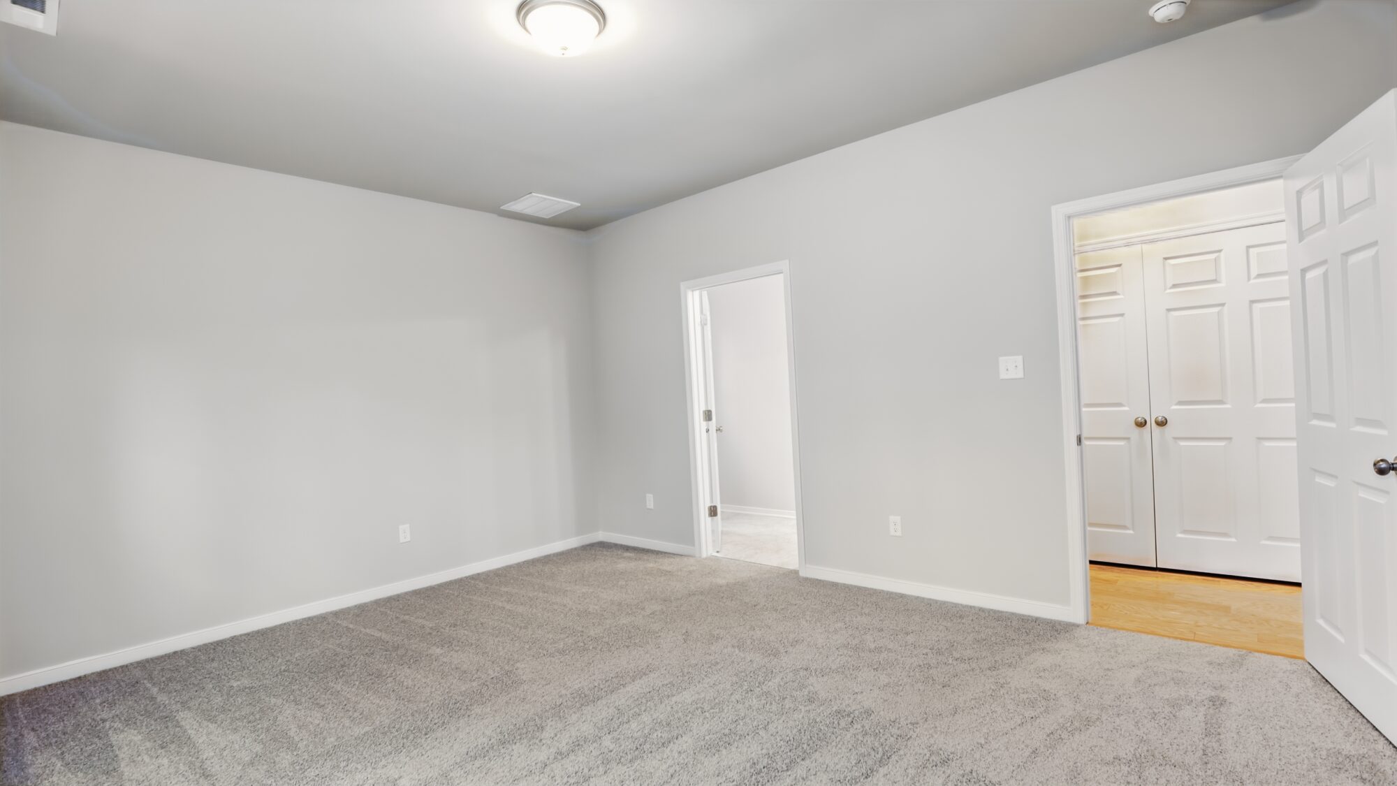Empty room at Somerset Hill with light gray walls, gray carpet, ceiling light, and open doors to other rooms.